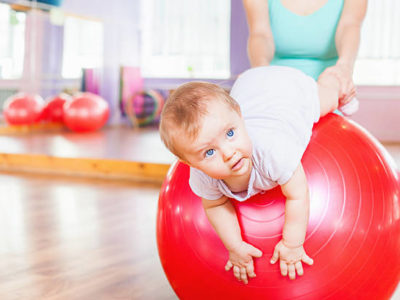 Mother with happy baby doing exercises with red gymnastic ball at fitness class. Concept of caring for the baby's health.
